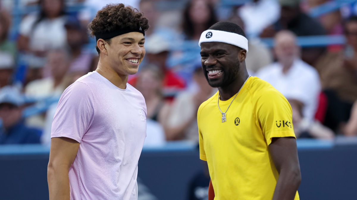 Two smiling tennis players stand together courtside, wearing headbands and contrasting lavender and yellow shirts.