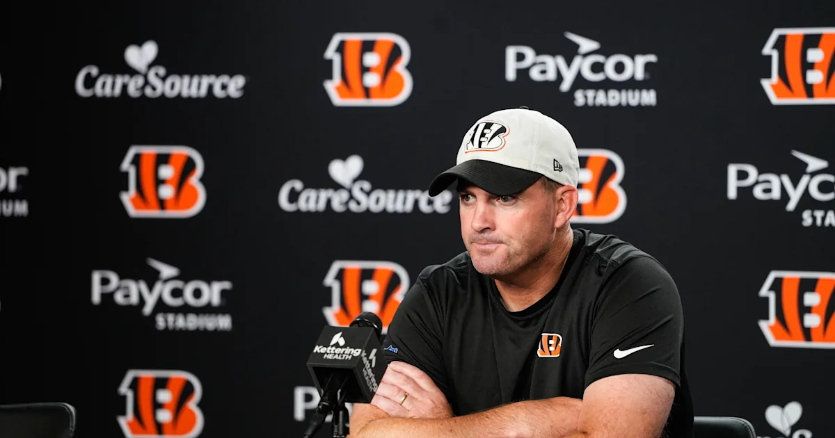 A man in a Bengals cap and black shirt sits at a press conference table before team sponsor logos.