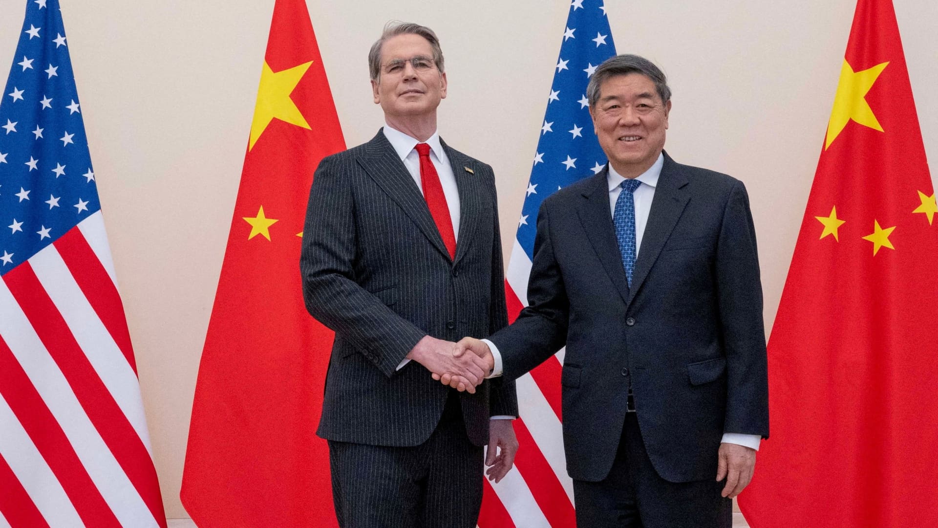 Two officials shake hands between American and Chinese flags during a formal diplomatic meeting.