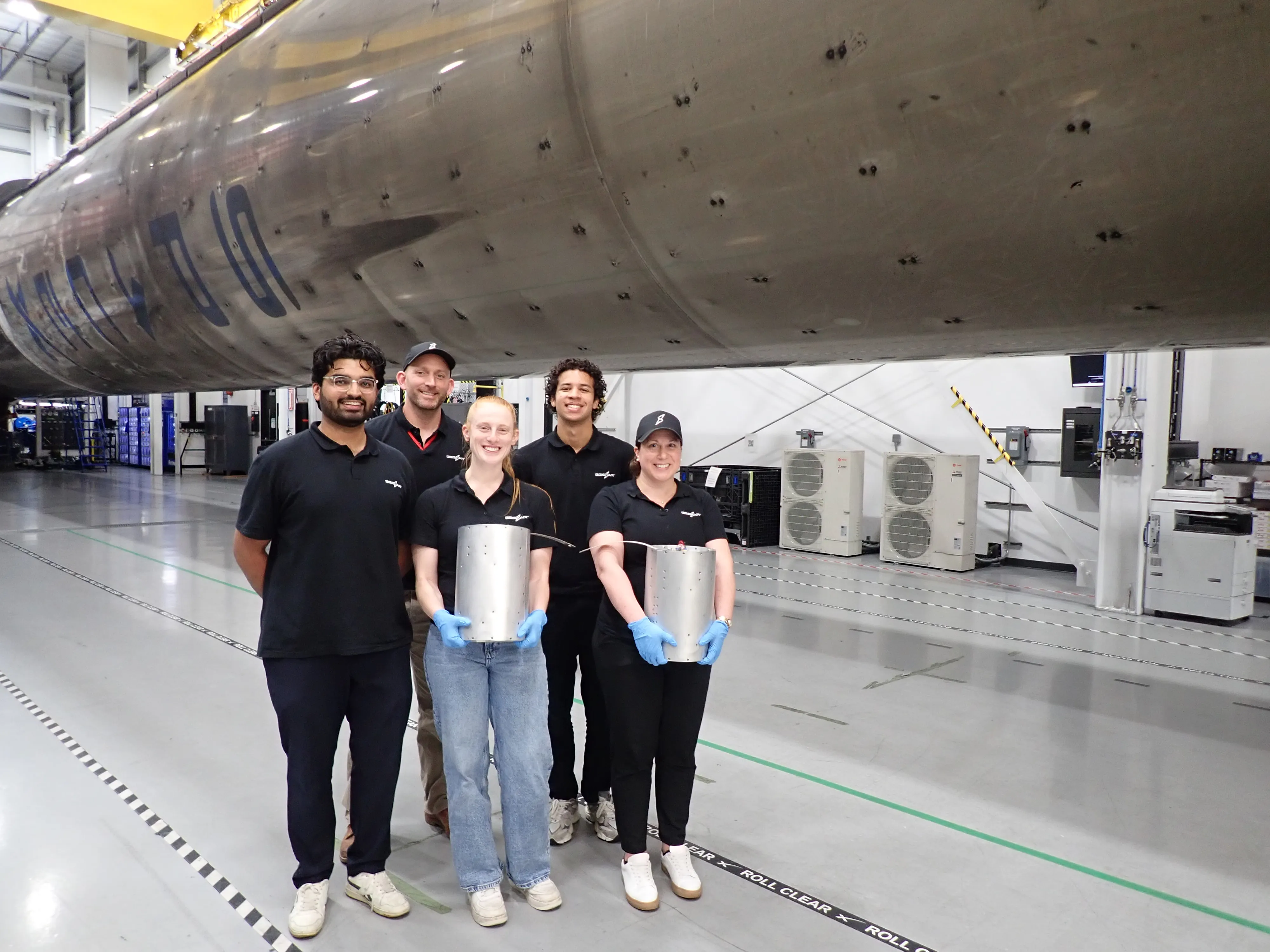 Five people pose beneath a large spacecraft structure inside an industrial facility, holding metal components.