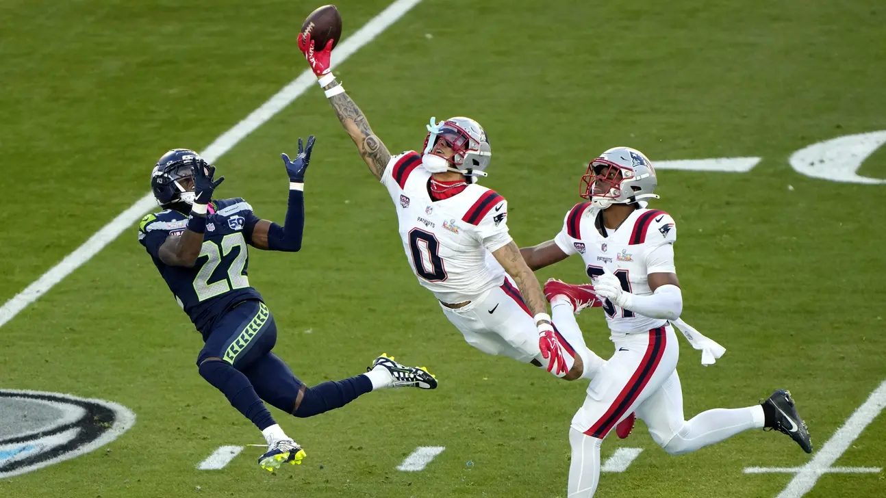 Football players from the Patriots and Seahawks leap and reach for a pass during play.