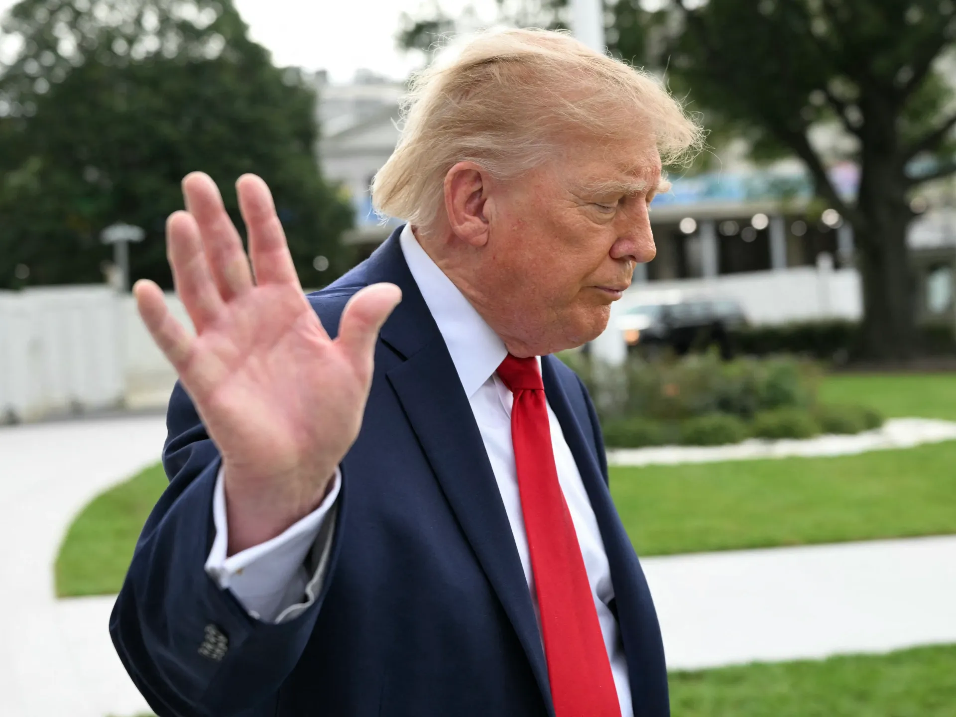 Donald Trump in a navy suit and red tie raises an open hand outdoors.