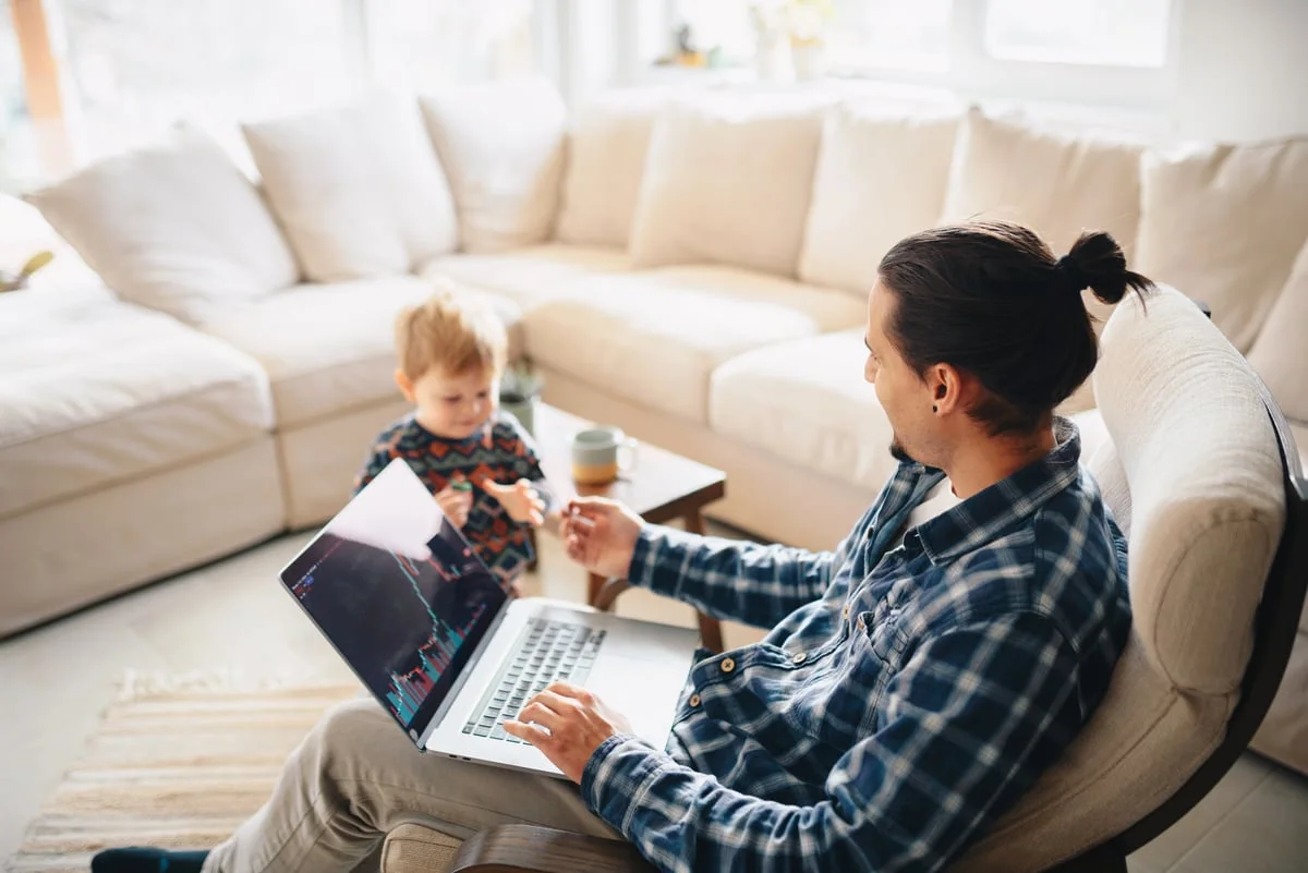 Man using a laptop beside a young child in a bright living room.