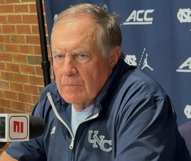 Older man in UNC jacket speaking into a microphone during a press conference