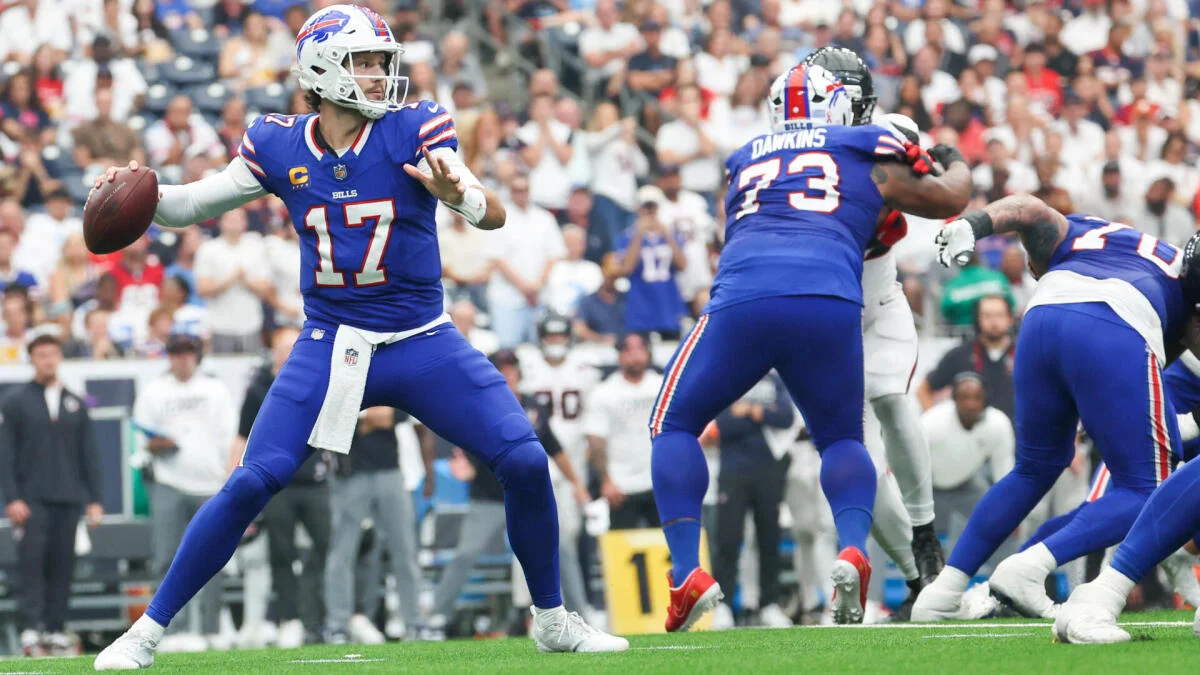 Buffalo Bills quarterback in blue uniform prepares to throw as teammates block defenders during a football game.