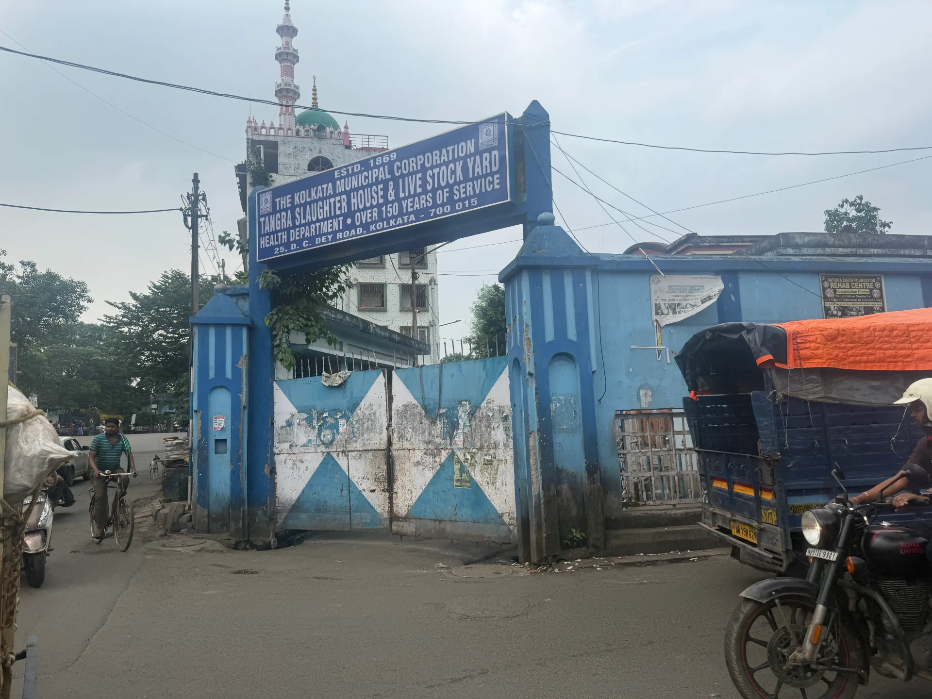 Street entrance with blue gate, slaughterhouse sign, mosque minaret, bicycle, motorcycles and covered handcart