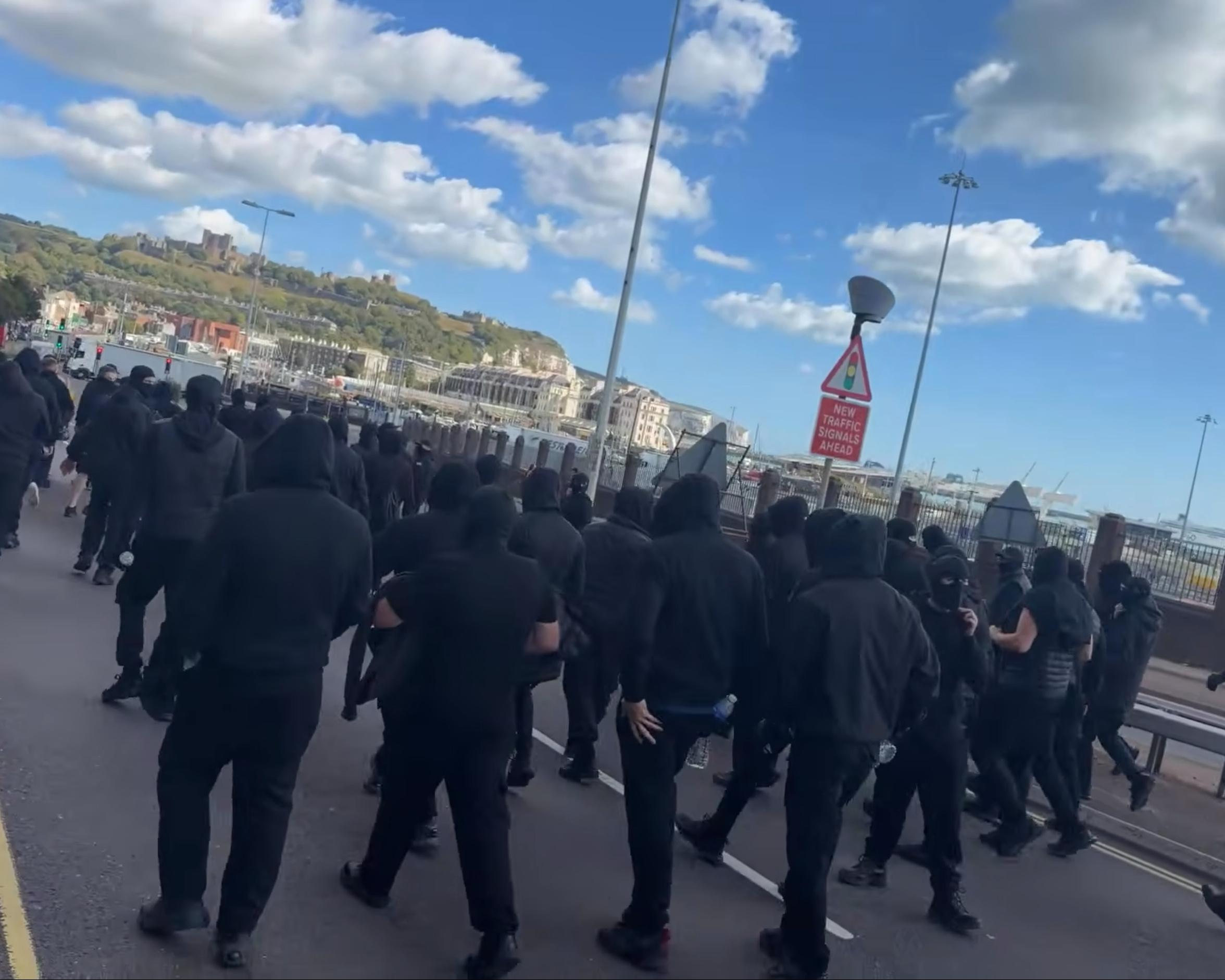 Crowd of black-clad people walking along a waterfront roadway beneath a bright blue sky