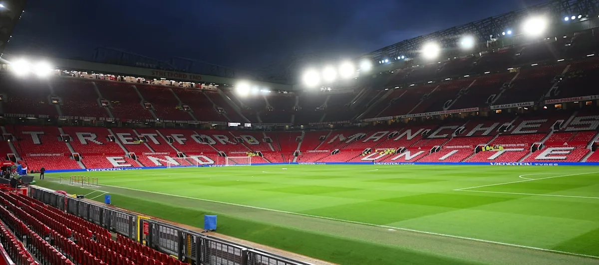Floodlit football stadium with empty red seats, green pitch, and Manchester United lettering across the stands