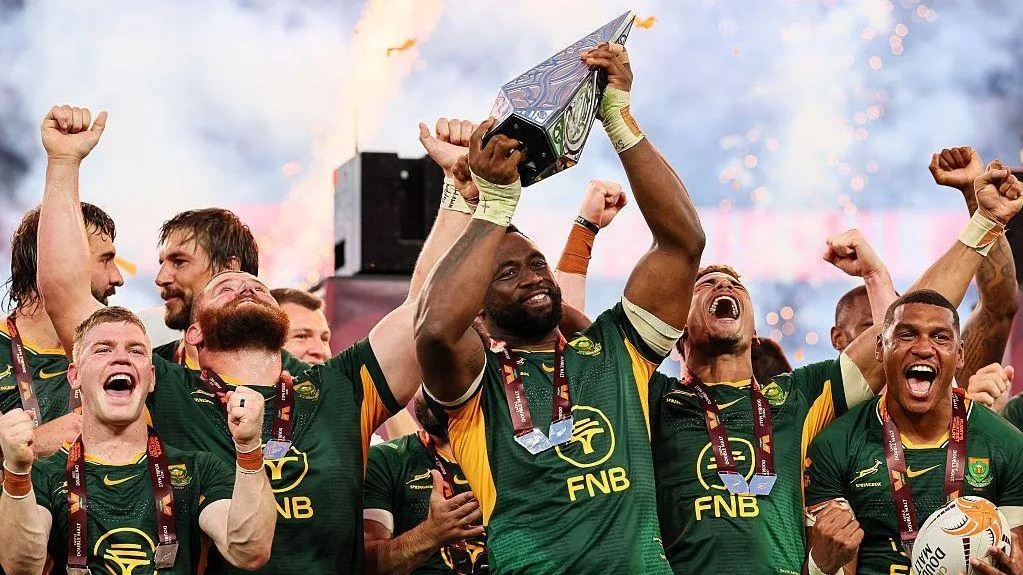 Rugby players in green jerseys celebrate while lifting a trophy amid fireworks.