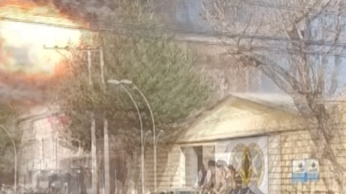 Blurry street scene showing a damaged building, trees, and indistinct people near its entrance.