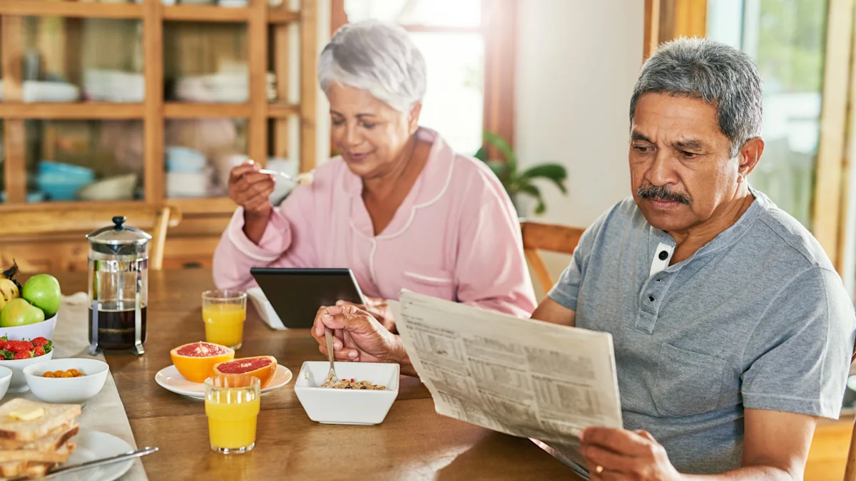 Older couple having breakfast together, with one reading a newspaper and the other using a tablet.