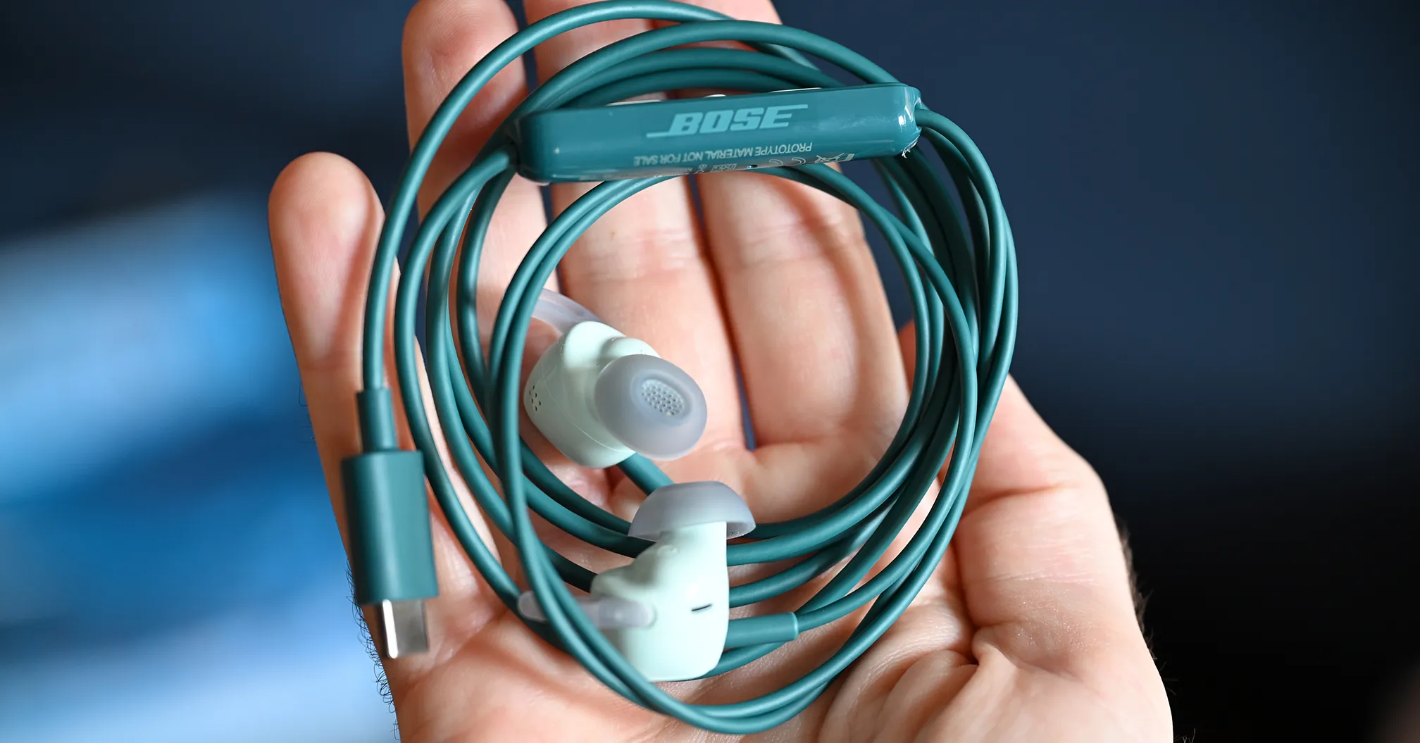 A hand holds teal wired earbuds with a USB-C plug and Bose inline remote.