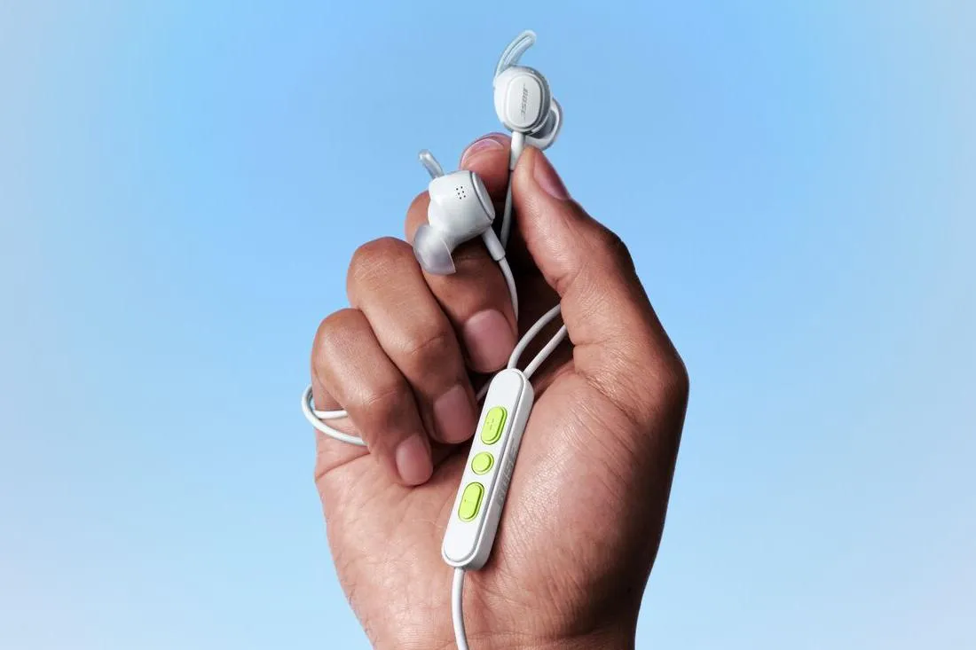 A hand holds white wired earbuds with lime-green inline controls against a light blue background.