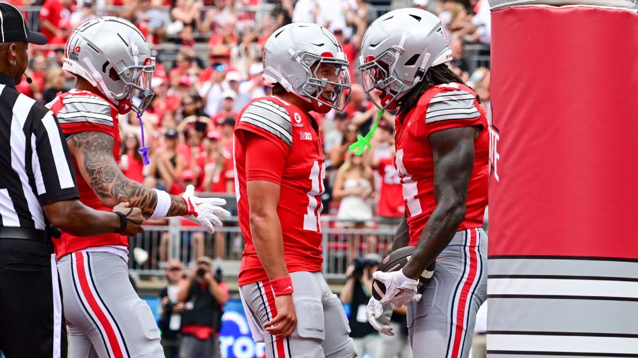 Three football players in red uniforms stand together near a sideline official during a game.