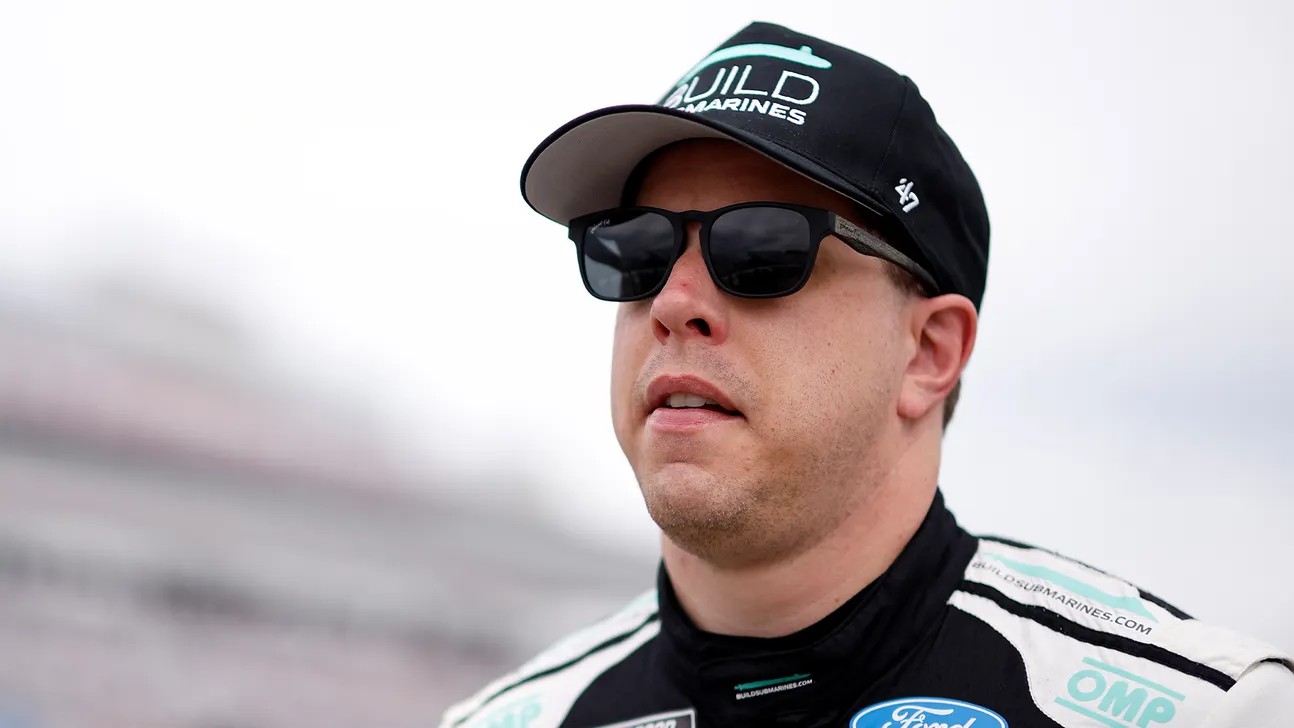 A man in a black racing cap, dark sunglasses, and racing suit looks ahead.