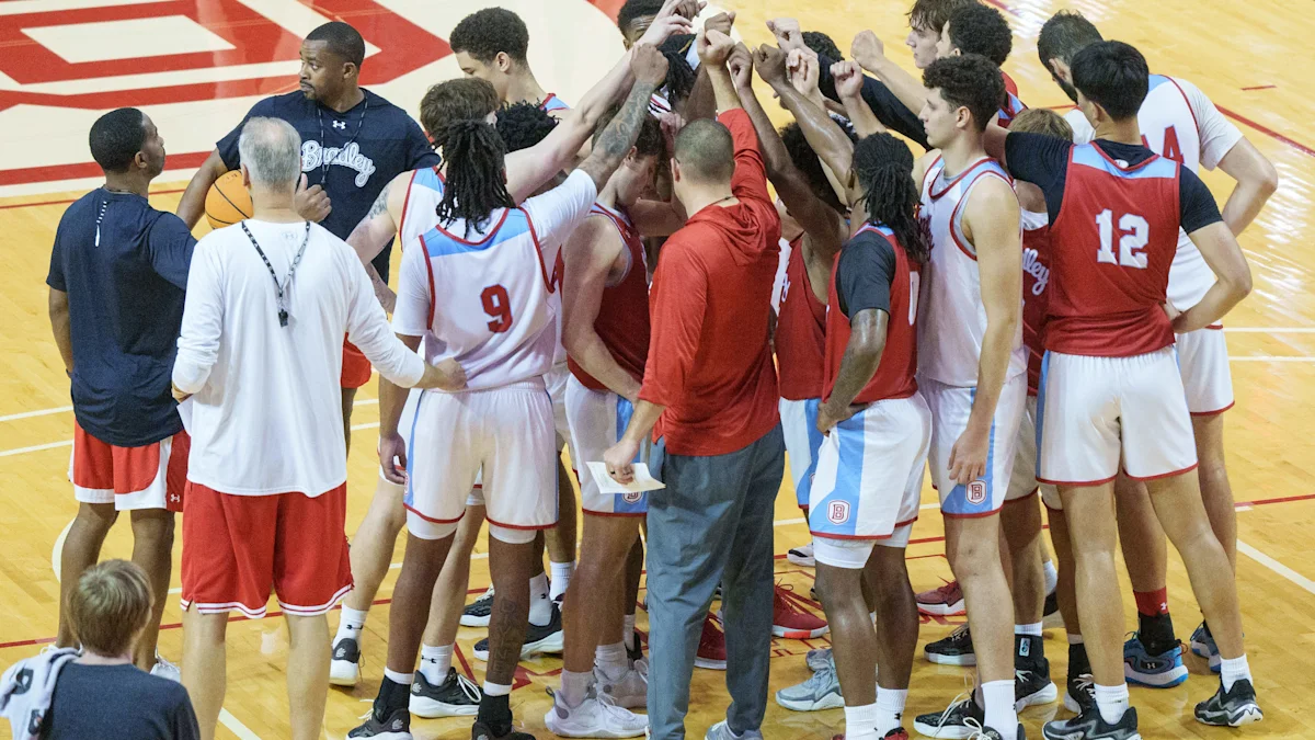 Basketball players and coaches gather with raised hands in a gym before a team huddle.