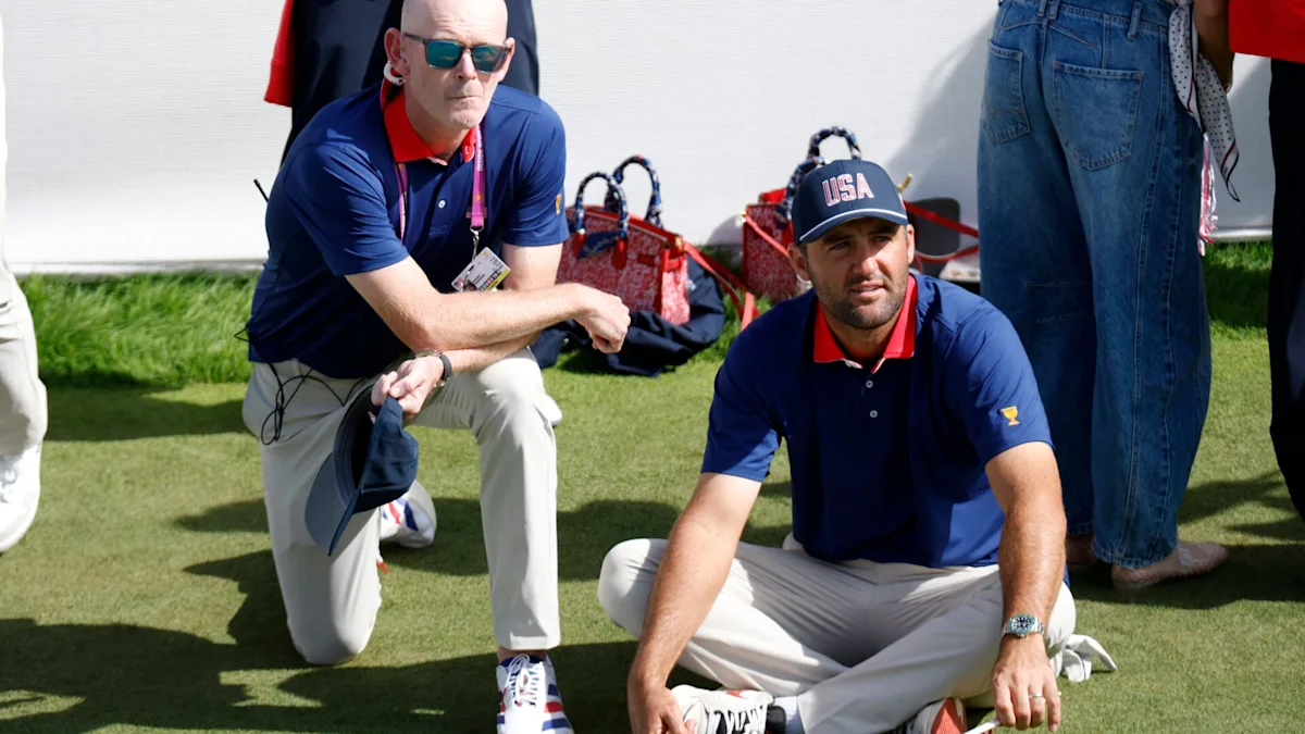 Two men in navy golf team shirts sit on grass beside equipment and other people.