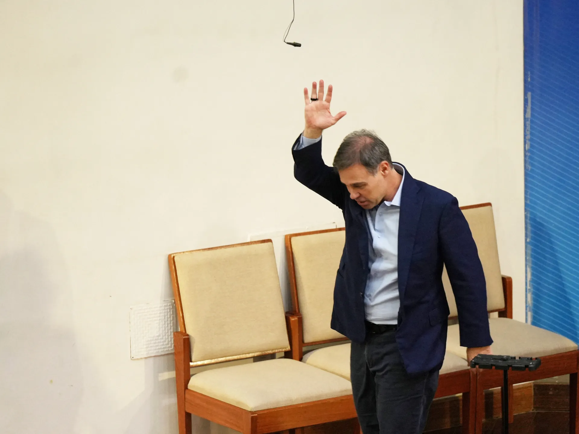 Man in a dark suit raises one hand while standing beside empty chairs and a lectern.