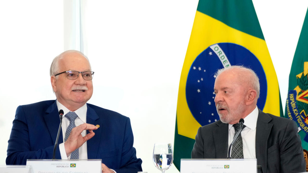 Two Brazilian officials speak at a table before Brazilian flags during a formal event