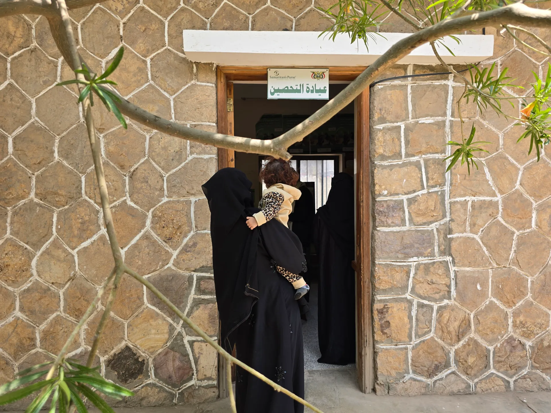 Women in black garments carry a child through a stone clinic doorway beneath an Arabic vaccination sign.