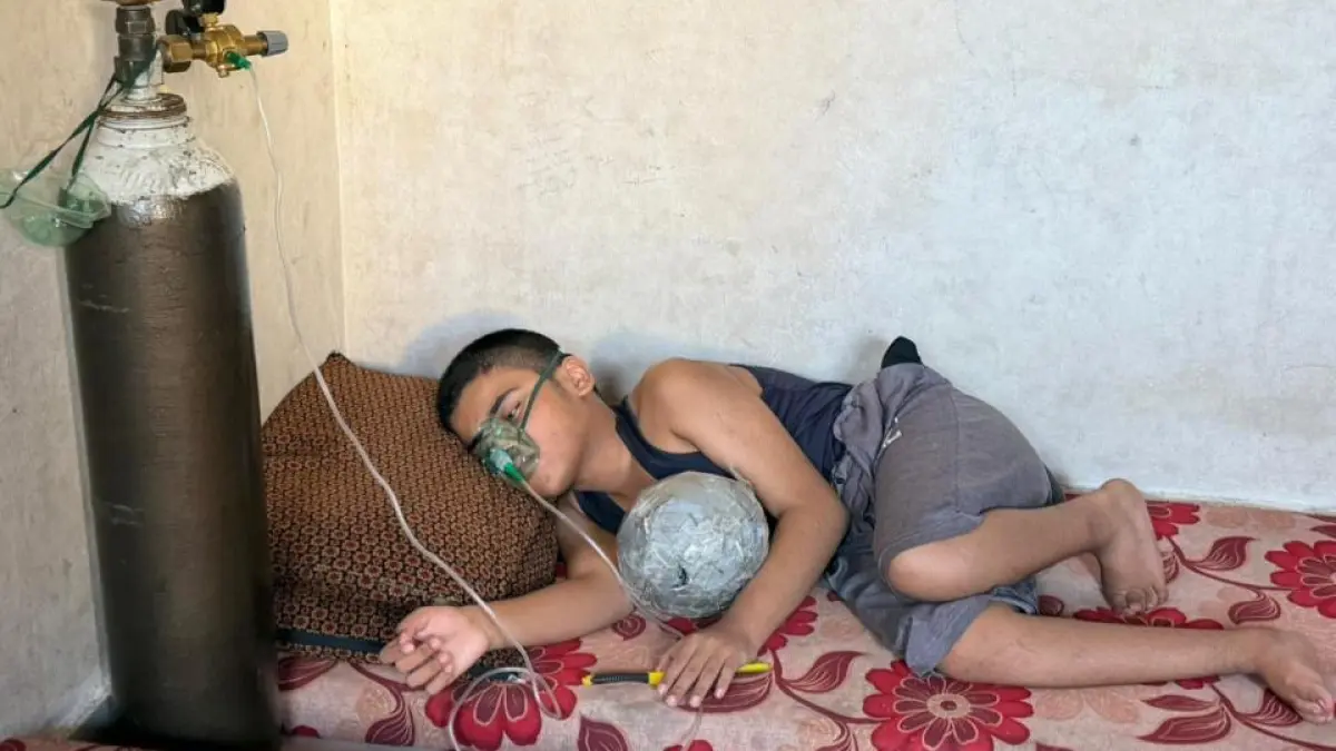 A boy lies on a bed wearing an oxygen mask beside a large cylinder.