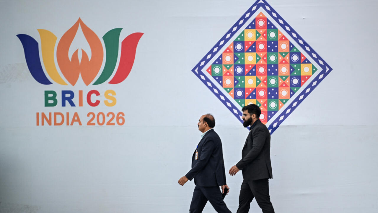 Two men walk past BRICS India 2026 branding and a colorful geometric emblem.