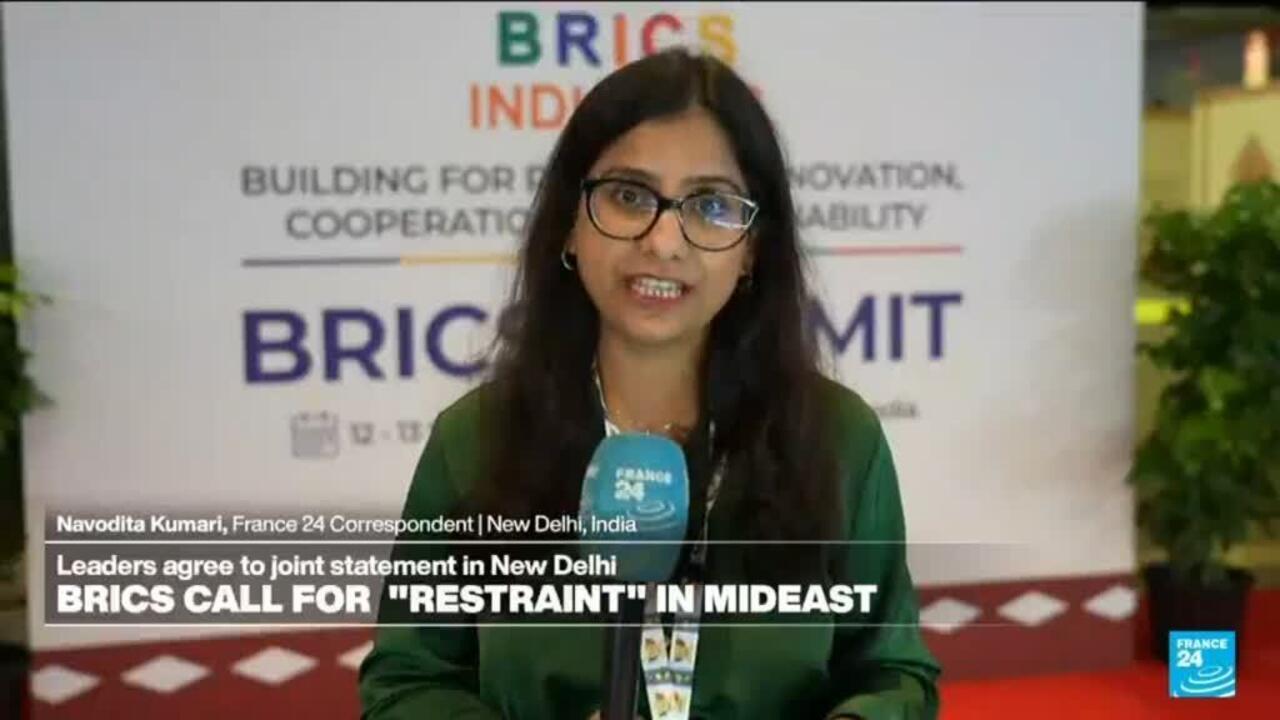 France 24 correspondent speaks before a BRICS summit backdrop, holding a blue microphone.