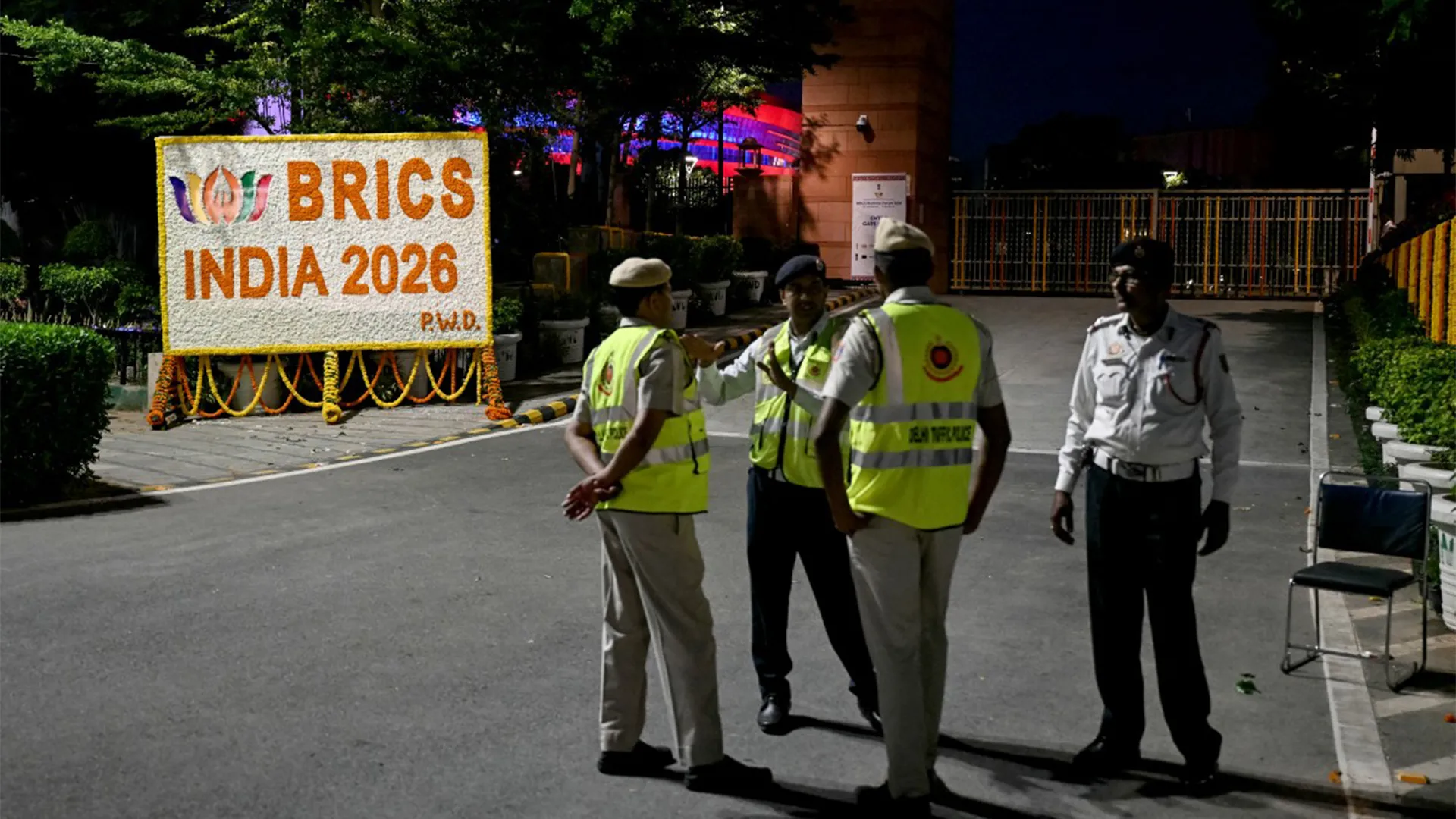 Security personnel stand beside a floral BRICS India 2026 sign outside a guarded venue at night.