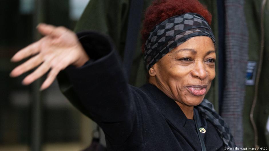 A smiling woman in a patterned headwrap raises her hand outdoors.