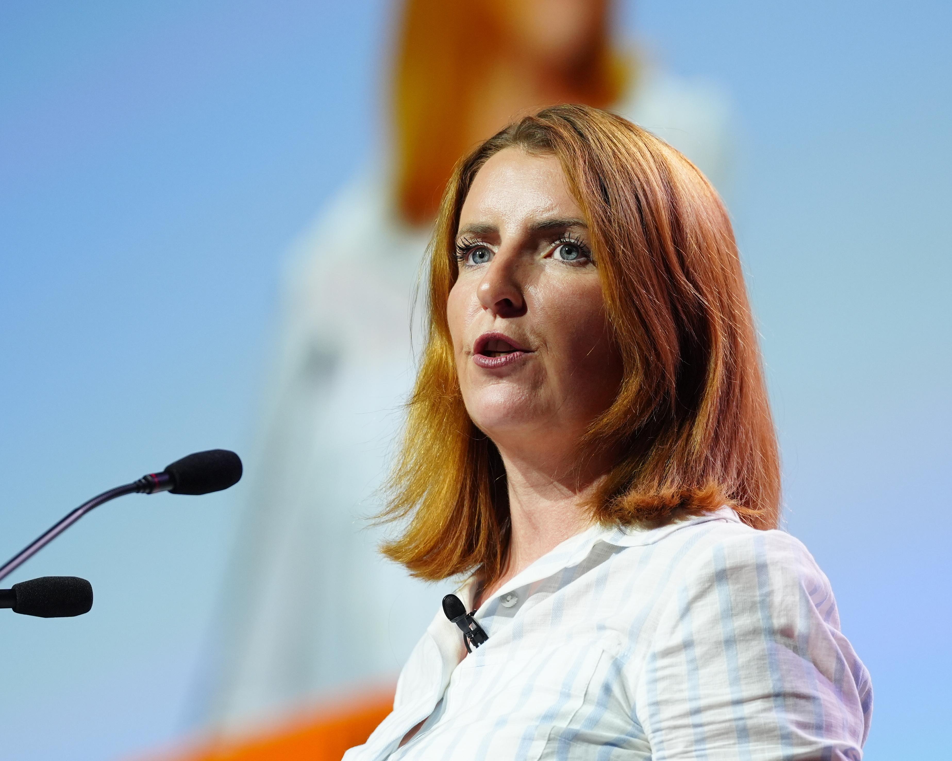 Red-haired woman speaking at a podium microphone against a pale blue background