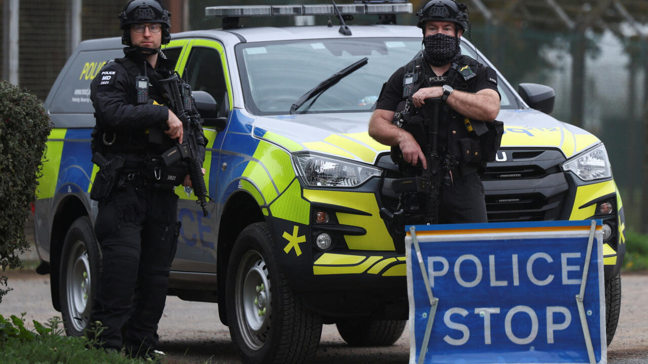 Two armed police officers stand in front of a marked police vehicle and a police stop sign.