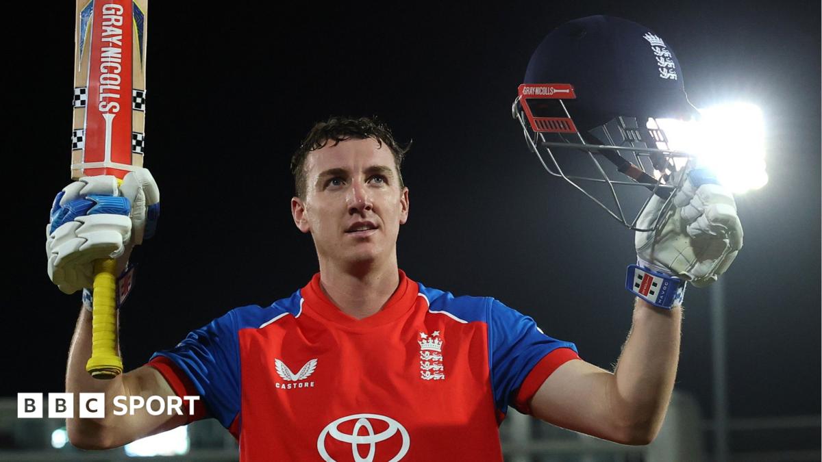Cricketer in red and blue England shirt raises bat and helmet under bright stadium lights