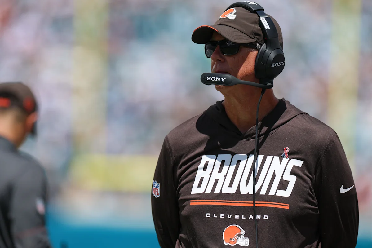 Cleveland Browns coach wearing a headset, sunglasses, cap, and team sweatshirt on the sideline
