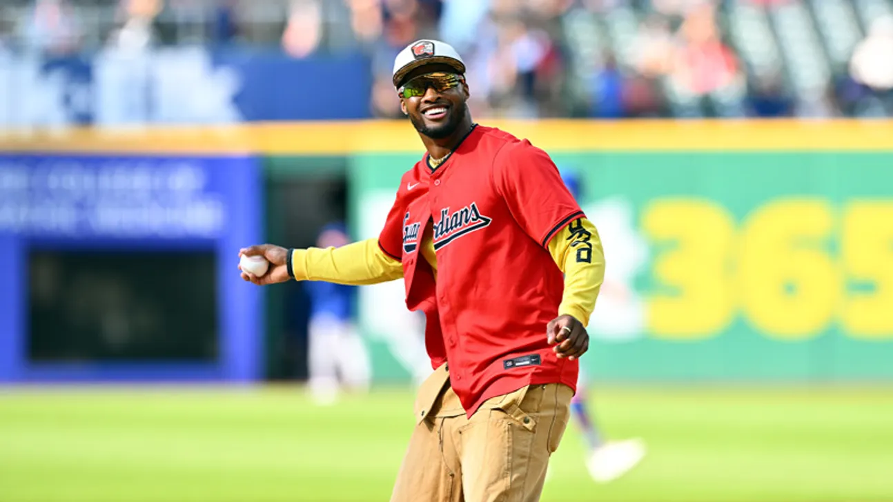 Smiling baseball player in a red Guardians jersey holds a ball on the field.