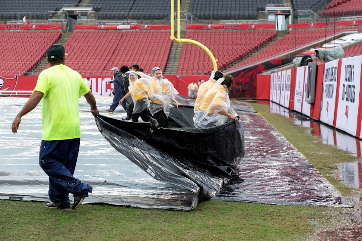 Workers pull a large tarp across a rain-soaked football field as heavy rain falls.
