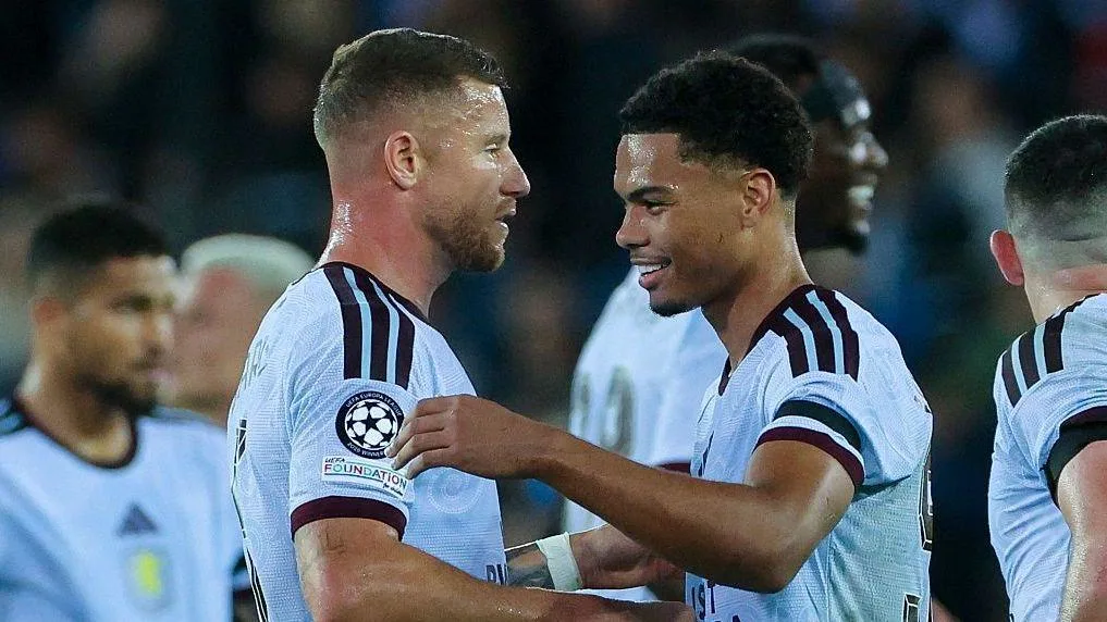Two footballers in white kits smile and talk during a match, with teammates and spectators behind them.