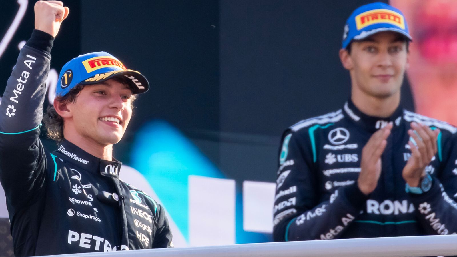 Two Formula 1 drivers in racing suits celebrate on a podium, one raising his fist and another applauding.
