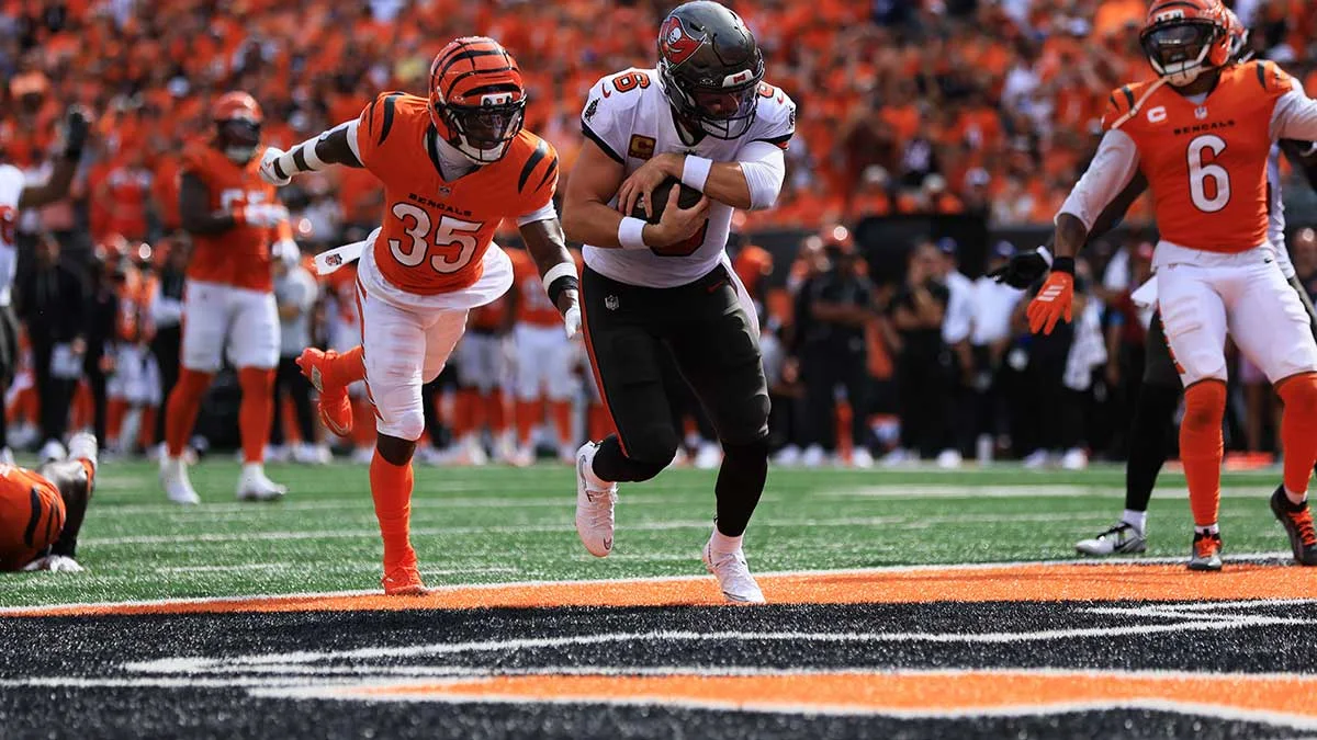 Football player in white jersey runs with ball as defenders in orange chase across end zone.