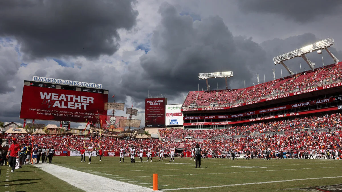 Football players stand on a field beneath storm clouds inside a crowded stadium with weather alerts displayed.