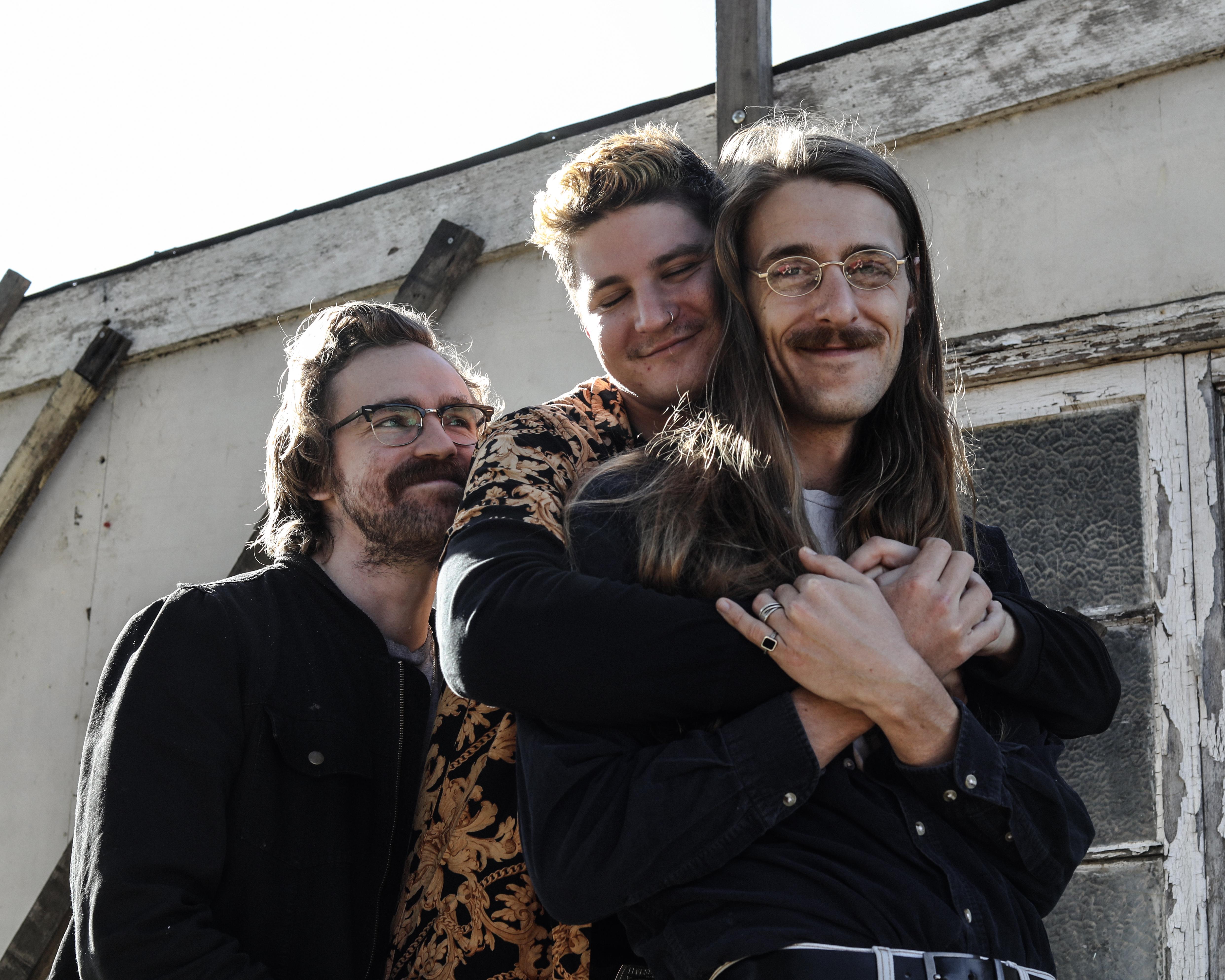 Three men embrace outside a weathered building in bright sunlight