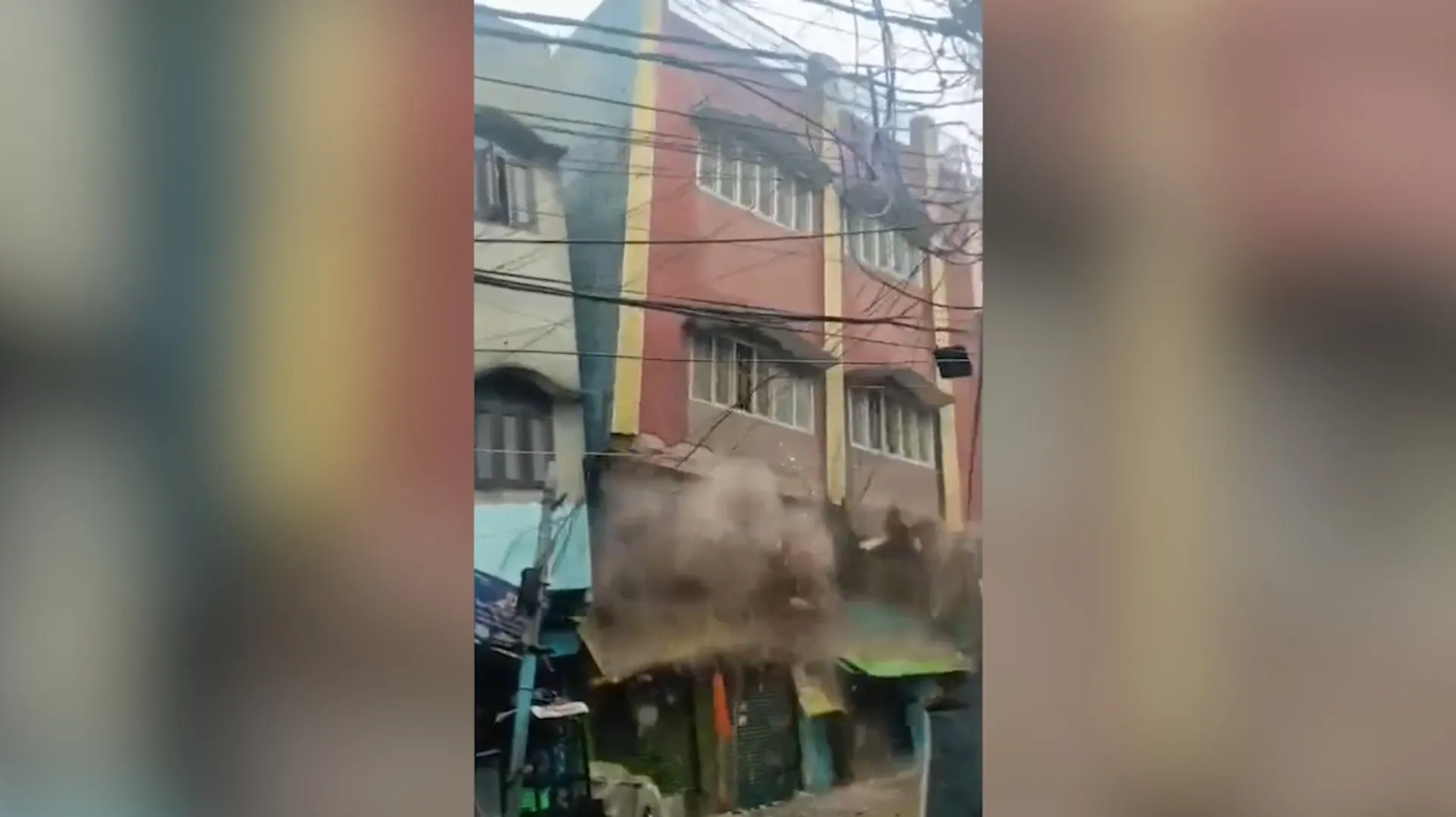 Dust billows from a partially collapsed multistory building beside tangled overhead wires.