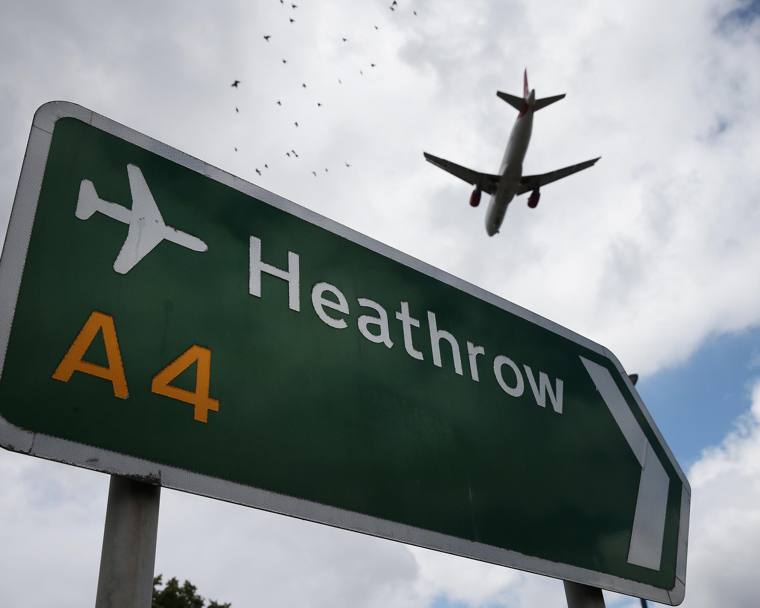 A Heathrow airport road sign fills the foreground as a passenger plane flies overhead beneath a cloudy sky.