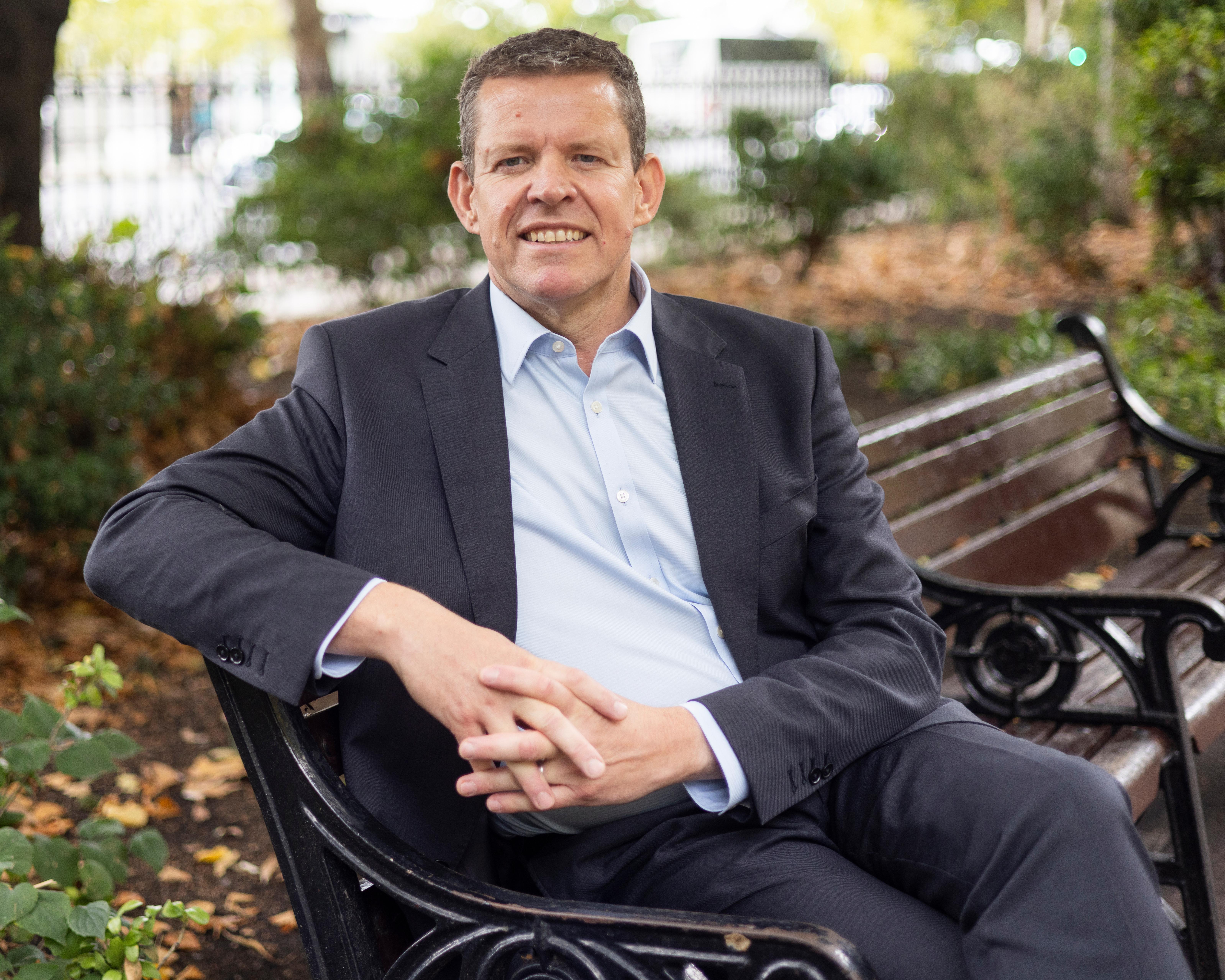 Man in a dark suit sitting on a park bench with hands clasped
