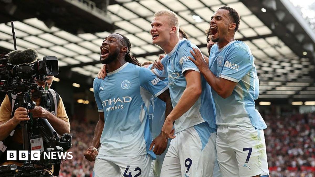 Manchester City players celebrate together on the pitch beside a television camera operator.