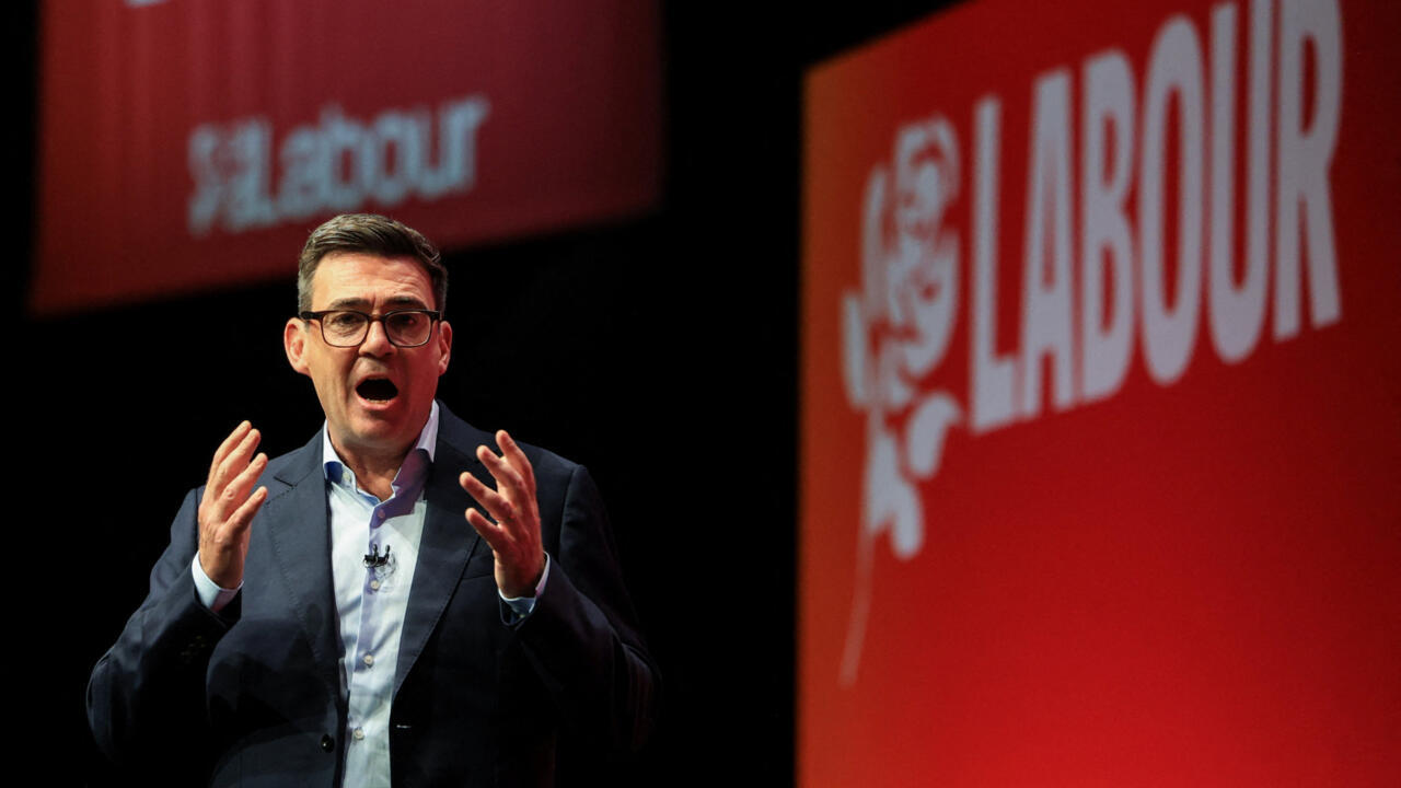 A man in glasses speaks with raised hands beside a large red Labour Party sign.