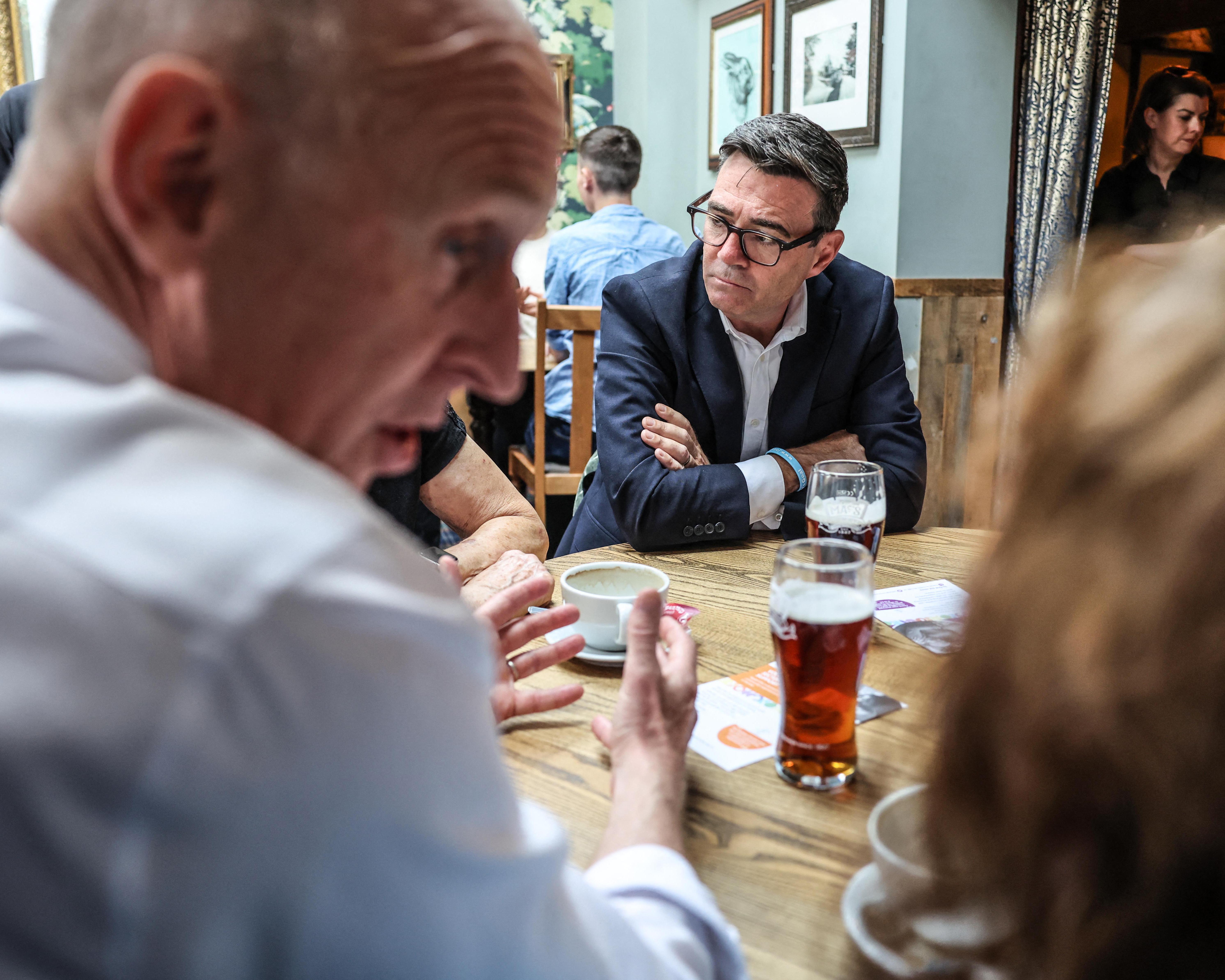 People converse around a pub table as a bespectacled man in a suit listens.