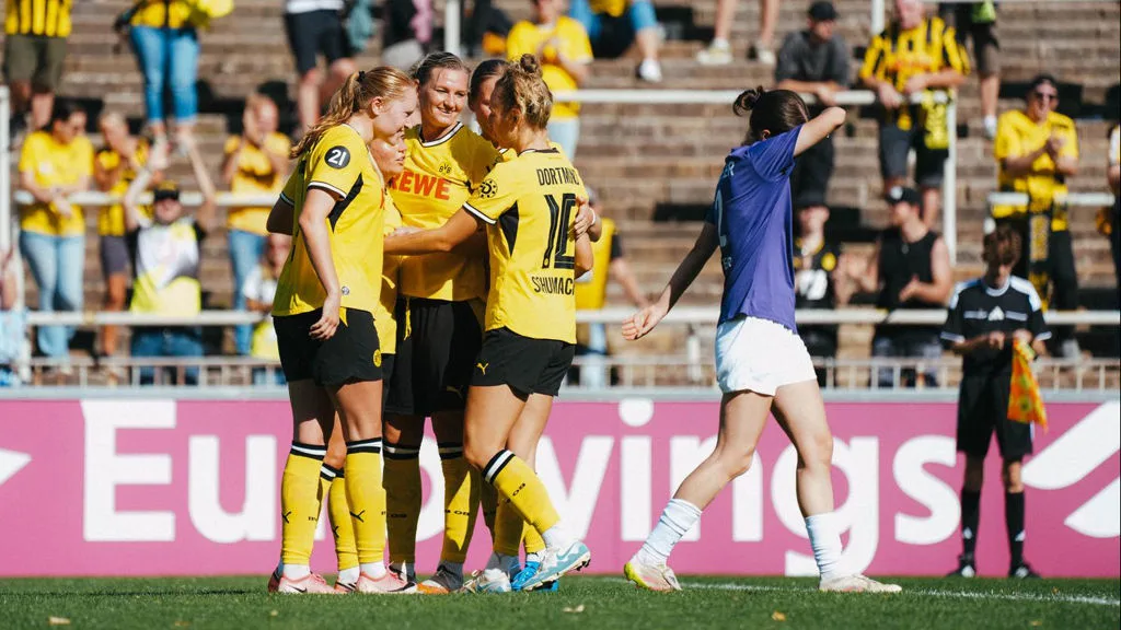 Dortmund women’s football players in yellow jerseys celebrate as an opposing player walks past.