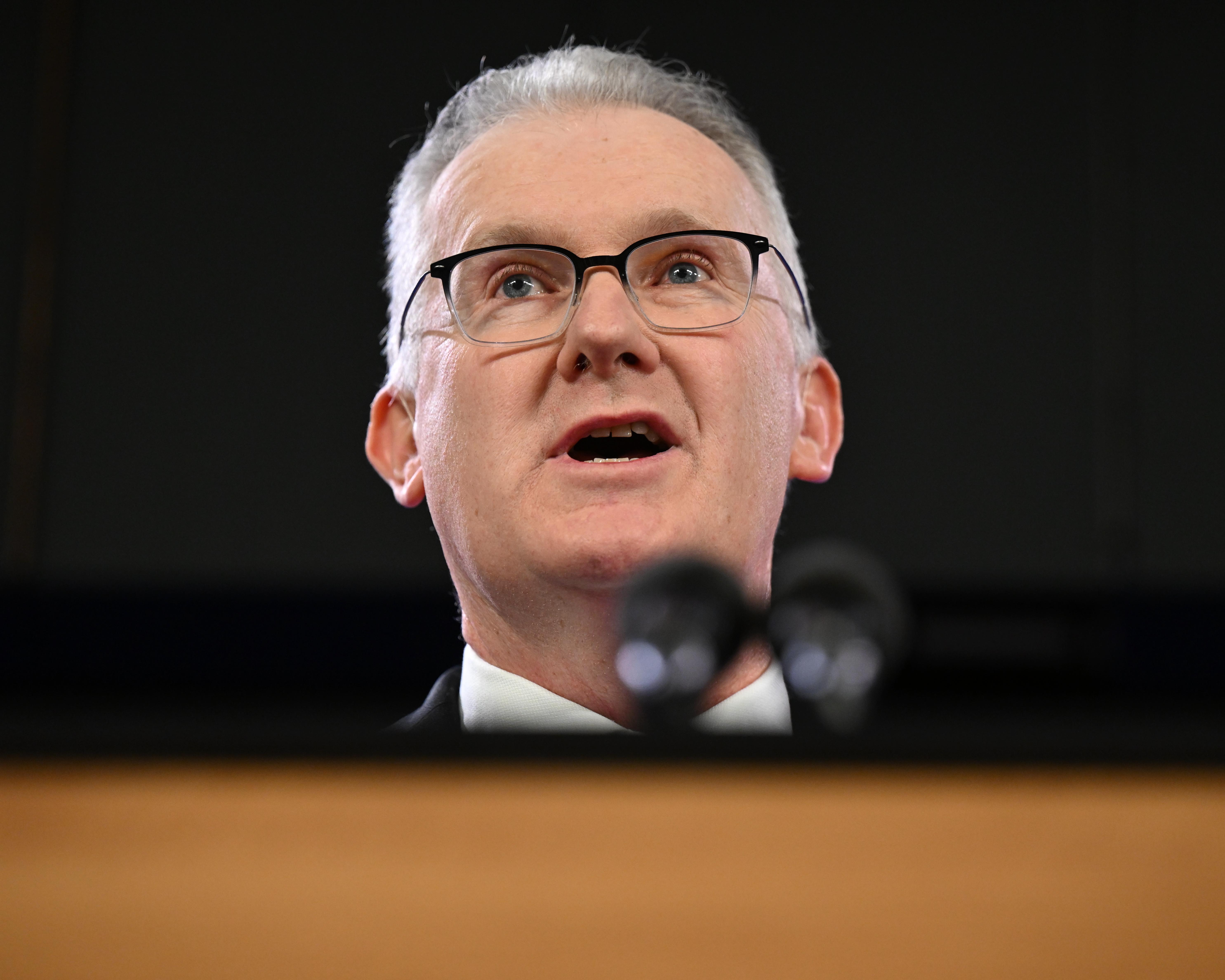 White-haired man wearing glasses speaks at a hearing, with microphones in the foreground.