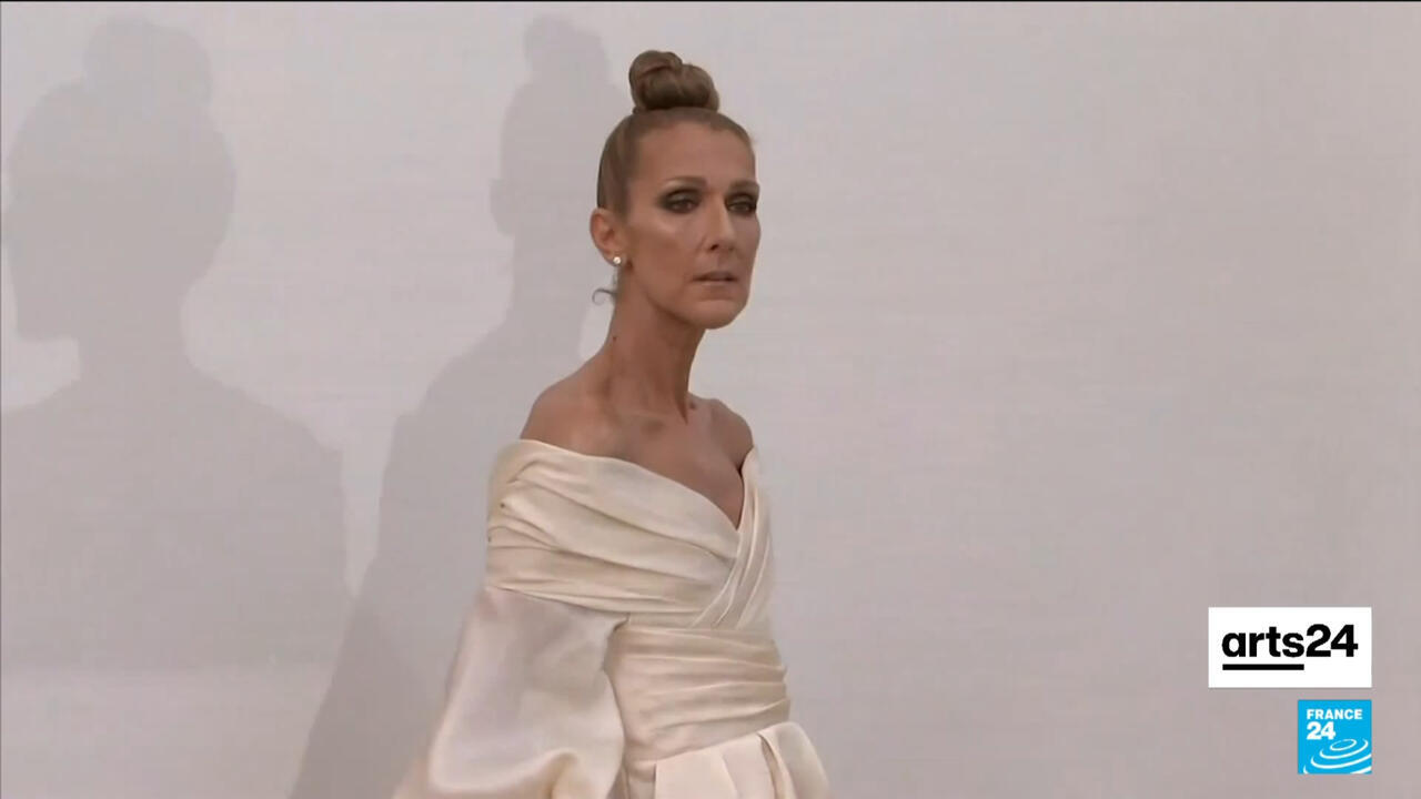 Woman in an ivory off-the-shoulder gown posing against a pale wall with shadows