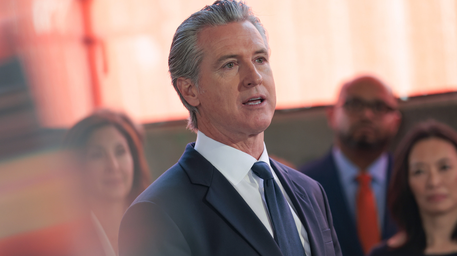 Gray-haired man in a dark suit speaks before a softly focused group of people.
